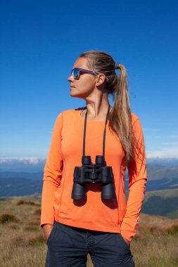 Göğsünde dürbün olan güneş gözlüklü kız dağlardaki mesafeye bakıyor.