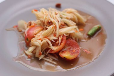 Tabakta Tayland papayası salatası, kakalı papaya salatası, limon, biber, beyaz bir masada gri bir tabakta kırmızı domates..