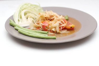 Tabakta Tayland papayası salatası, kakalı papaya salatası, limon, biber, beyaz bir masada gri bir tabakta kırmızı domates..