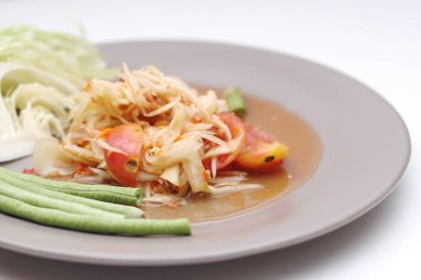 Tabakta Tayland papayası salatası, kakalı papaya salatası, limon, biber, beyaz bir masada gri bir tabakta kırmızı domates..