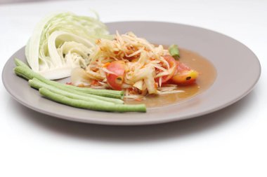 Tabakta Tayland papayası salatası, kakalı papaya salatası, limon, biber, beyaz bir masada gri bir tabakta kırmızı domates..