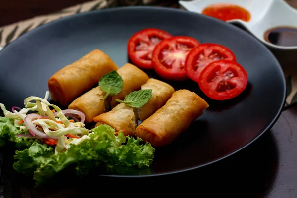 Fried spring rolls with vegetables and tomatoes placed in a black plate on a black wooden table and dipping sauce in tjhailand.