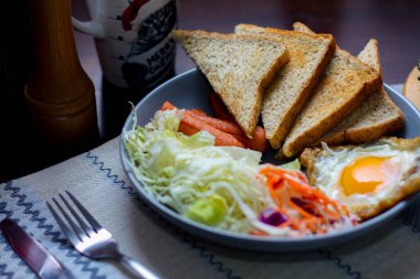 Kahvaltı, sahanda yumurta, kızarmış sosis, sebze salatası ve kahverengi ahşap bir masada tost ve Tayland kahvesi..