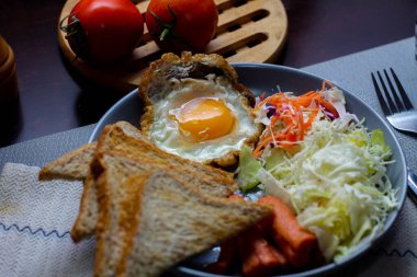 Kahvaltı, sahanda yumurta, kızarmış sosis, sebze salatası ve kahverengi ahşap bir masada tost ve Tayland kahvesi..