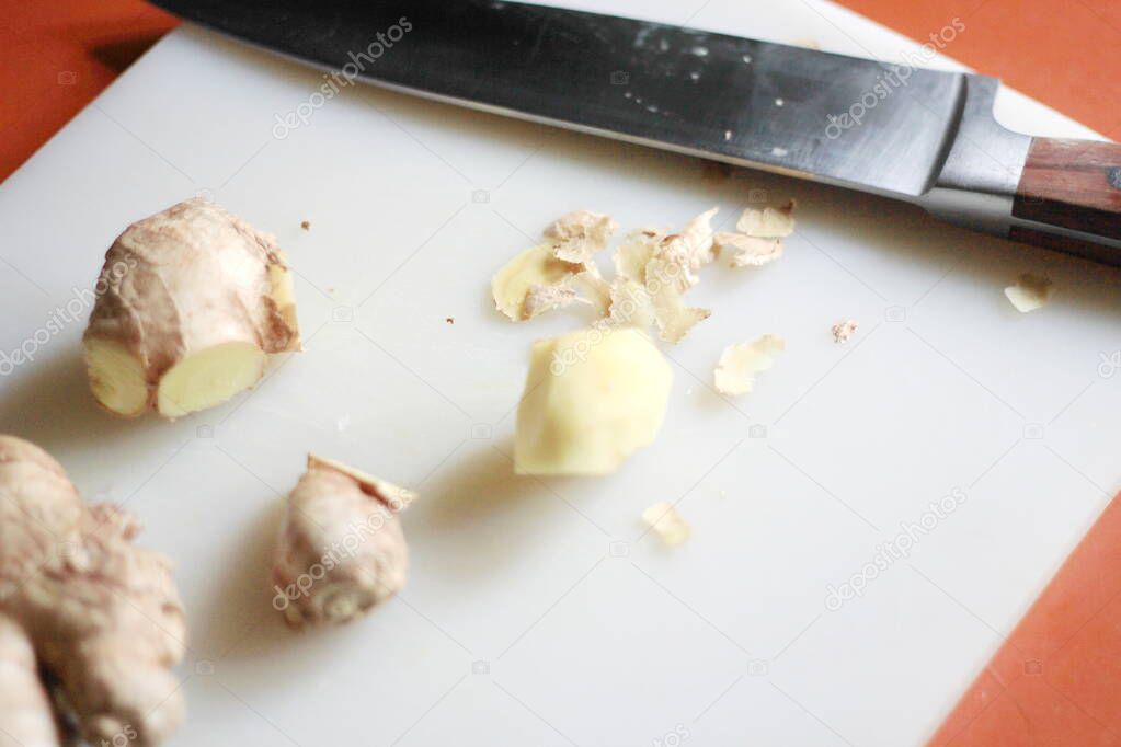 Ginger is being sliced on a plastic chopping board with a knife on an orange table.