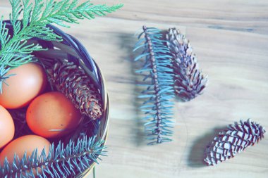 Çam yaprakları, kozalaklar ve açık kahverengi ahşap zemine yerleştirilmiş dallarla dolu bir sepette yumurtalar..