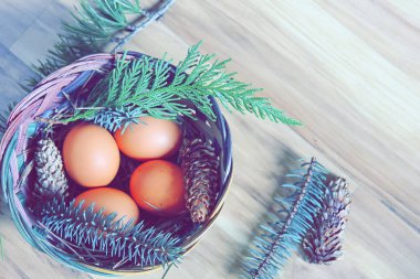 Çam yaprakları, kozalaklar ve açık kahverengi ahşap zemine yerleştirilmiş dallarla dolu bir sepette yumurtalar..