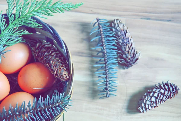 Çam yaprakları, kozalaklar ve açık kahverengi ahşap zemine yerleştirilmiş dallarla dolu bir sepette yumurtalar..