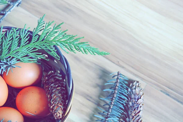 Çam yaprakları, kozalaklar ve açık kahverengi ahşap zemine yerleştirilmiş dallarla dolu bir sepette yumurtalar..