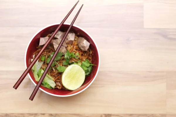 Stewed pork Noodle garnished with coriander, sliced spring onions and lemon wedges. Served on an ash wood table