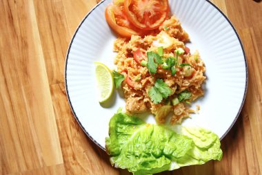 Domuz eti ve kızarmış pilav hafif ahşap bir masada beyaz bir tabakta servis edilir. Limonlu, sebzeli ve domatesli açık ahşap bir masa..