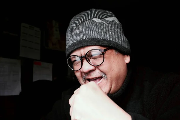 Bearded Asian man in black jacket and knitted hat. Working in a dark room and looking happy at the computer