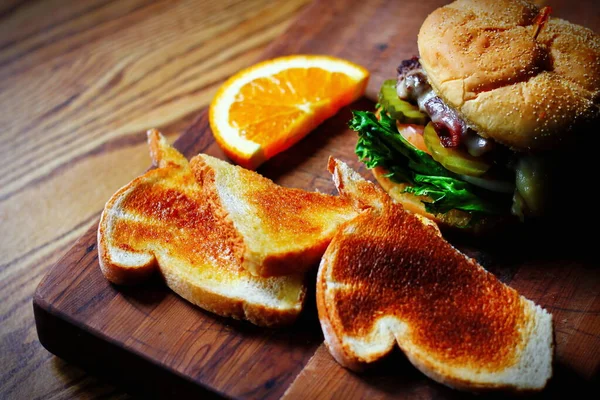 Photo of deluxe burger, hamburger being decorated before serving on a wooden chopping board consisting of toast and orange slices. Burgers are popular food in America.