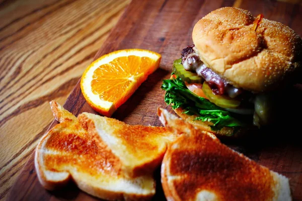 Photo of deluxe burger, hamburger being decorated before serving on a wooden chopping board consisting of toast and orange slices. Burgers are popular food in America.