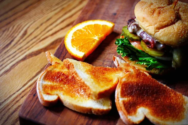 Photo of deluxe burger, hamburger being decorated before serving on a wooden chopping board consisting of toast and orange slices. Burgers are popular food in America.