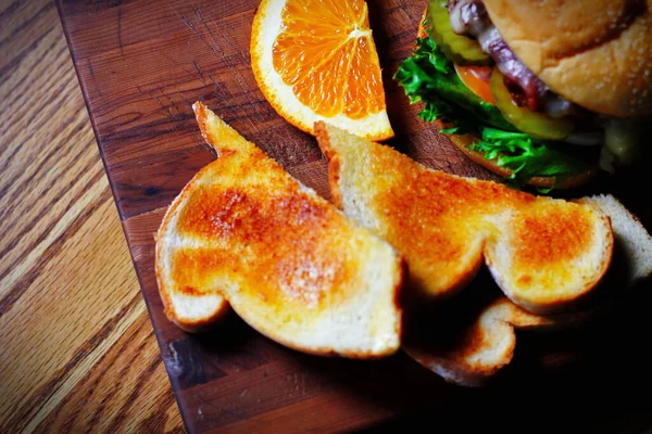 Photo of deluxe burger, hamburger being decorated before serving on a wooden chopping board consisting of toast and orange slices. Burgers are popular food in America.