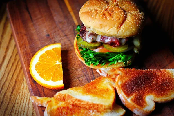 Photo of deluxe burger, hamburger being decorated before serving on a wooden chopping board consisting of toast and orange slices. Burgers are popular food in America.