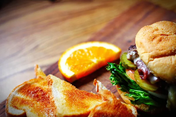 Photo of deluxe burger, hamburger being decorated before serving on a wooden chopping board consisting of toast and orange slices. Burgers are popular food in America.