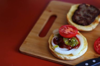 Hamburger dilimlenmiş domates, soğan ve turşuyla servis ediliyor. Mutfaktaki tahta kesme tahtasının üzerinde.