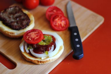 Hamburger dilimlenmiş domates, soğan ve turşuyla servis ediliyor. Mutfaktaki tahta kesme tahtasının üzerinde.