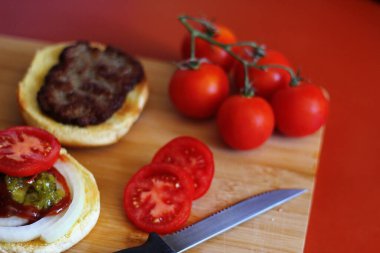 Hamburger dilimlenmiş domates, soğan ve turşuyla servis ediliyor. Mutfaktaki tahta kesme tahtasının üzerinde.