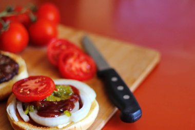 Hamburger dilimlenmiş domates, soğan ve turşuyla servis ediliyor. Mutfaktaki tahta kesme tahtasının üzerinde.