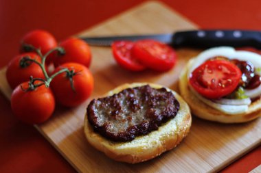 Hamburger dilimlenmiş domates, soğan ve turşuyla servis ediliyor. Mutfaktaki tahta kesme tahtasının üzerinde.