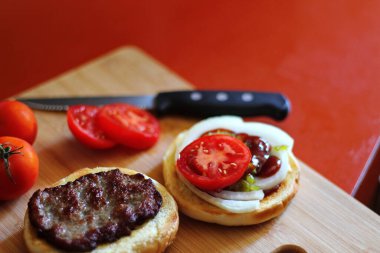 Hamburger dilimlenmiş domates, soğan ve turşuyla servis ediliyor. Mutfaktaki tahta kesme tahtasının üzerinde.
