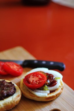 Hamburger dilimlenmiş domates, soğan ve turşuyla servis ediliyor. Mutfaktaki tahta kesme tahtasının üzerinde.