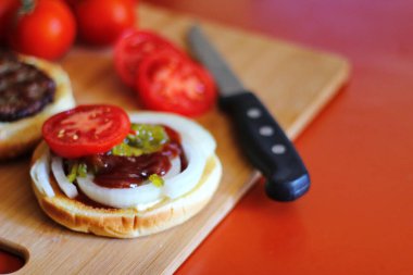 Hamburger dilimlenmiş domates, soğan ve turşuyla servis ediliyor. Mutfaktaki tahta kesme tahtasının üzerinde.