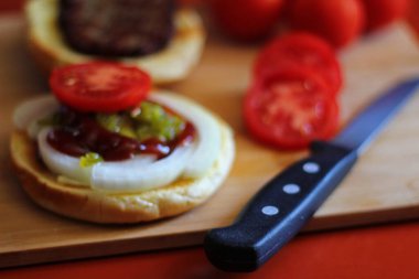 Hamburger dilimlenmiş domates, soğan ve turşuyla servis ediliyor. Mutfaktaki tahta kesme tahtasının üzerinde.
