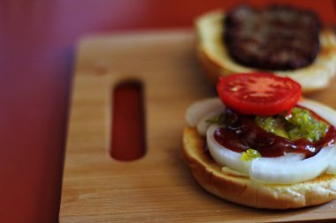 Hamburger dilimlenmiş domates, soğan ve turşuyla servis ediliyor. Mutfaktaki tahta kesme tahtasının üzerinde.