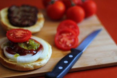 Hamburger dilimlenmiş domates, soğan ve turşuyla servis ediliyor. Mutfaktaki tahta kesme tahtasının üzerinde.