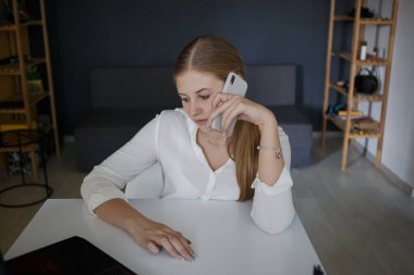 Beyaz tişörtlü, beyaz gömlekli genç ve başarılı bir iş kadını telefonda konuşuyor ve aşağı bakıyor, modern bir ofiste dizüstü bilgisayarla beyaz bir masa başında oturuyor. Kariyer başarısı, başarı ve kişisel büyüme kavramı