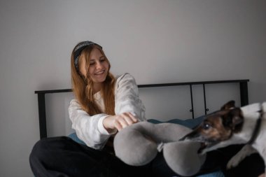 Kafasında bandajla ve uzun sarı saçlarıyla genç bir kız oturma odasında bir yatakta oturmuş köpekle oyun oynuyor ve gülümsüyor.