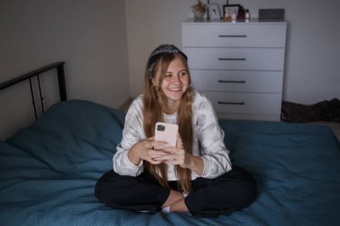 Kafasında bandajla ve uzun sarı saçlarıyla genç bir kız oturma odasında bir yatağa oturuyor, elinde bir telefon tutuyor ve gülüyor.