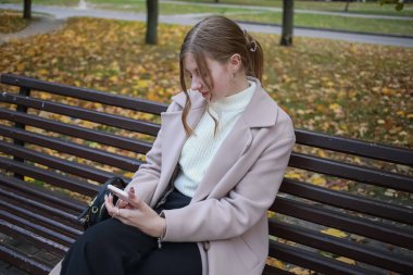 Güzel bir ceketi olan genç bir kız şehirde bir bankta oturuyor, saçları toplanmış, elinde bir telefon tutuyor ve gülümsüyor.