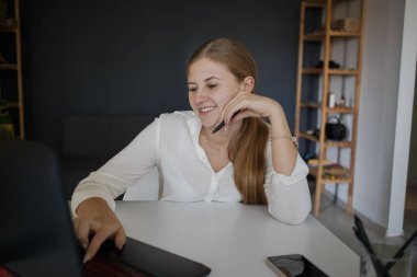 Beyaz gömlekli başarılı bir iş kadınının gülümseyen çalışanı ekrana bakıyor, elinde bir kalem tutuyor, iş yerinde oturuyor, modern bir ofiste dizüstü bilgisayarı olan beyaz bir masa. Kariyer başarısı, başarı...