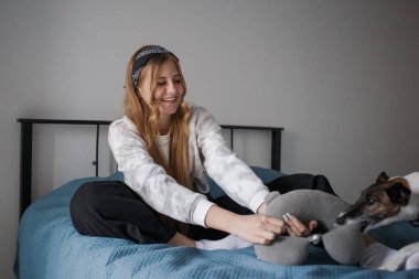 Kafasında bandajla ve uzun sarı saçlarıyla genç bir kız oturma odasında bir yatakta oturmuş köpekle oyun oynuyor ve gülümsüyor.