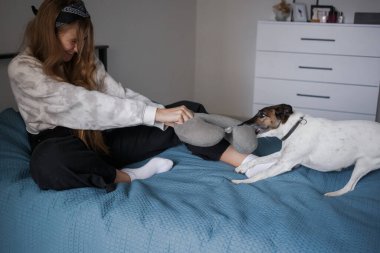 Kafasında bandajla ve uzun sarı saçlarıyla genç bir kız oturma odasında bir yatakta oturmuş bir köpekle oyun oynuyor.