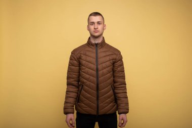 Stüdyo portresi. Sarı arka planda kahverengi ceketli genç bir adam üzgün duruyor.