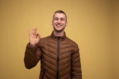 Stüdyo portresi. 20 'li yaşlarda, kahverengi ceketli, sarı arka planda, her şeyin yolunda olduğuna dair gülümseyen ve el kol hareketi yapan genç bir adam.