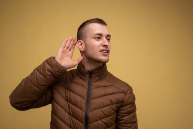 Kahverengi ceketli genç adam elini kulağına koyar ve dinler.