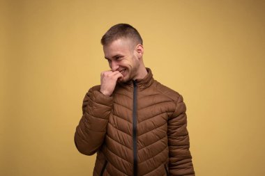 Stüdyo portresi. Sarı arka planda, kahverengi ceketli genç bir adam sinsice gülüyor yüzüne yumruk atıyor.
