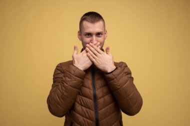 Stüdyo portresi. Sarı arka planda kahverengi ceketli genç bir adam, gülüyor ve elleriyle ağzını kapatıyor.