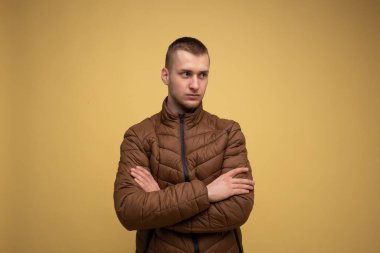 Stüdyo portresi. Sarı arka planda, kahverengi ceketli genç bir adam dikkatini dağıtmış ve kollarını göğsünde kavuşturmuş.