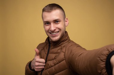 Stüdyo portresi. Sarı arka planda kahverengi ceketli genç bir adam cep telefonundan birinci şahıs selfie çekiyor ve sınıfını gösteriyor.