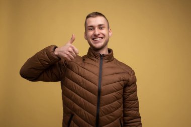 Stüdyo portresi. Sarı arka planda, kahverengi ceketli genç bir adam baş parmağını bir jest olarak gösteriyor.