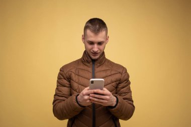 Stüdyo portresi. 20 'li yaşlarda, kahverengi ceketli, sarı arka planda, elinde cep telefonu olan genç bir adam cep telefonu kullanıyor, mesaj yazıyor, sohbet ediyor, SMS gönderiyor.