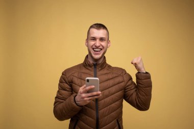 Stüdyo portresi. Kahverengi ceketli, sarı arka planda, elinde cep telefonu olan genç bir adam, kazanan bir jest yapar, yumruğunu sıkarak 